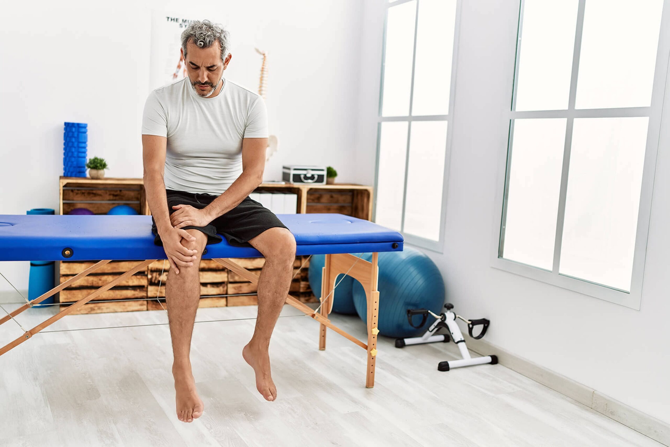 Man with curly, greying hair sits on a physical therapy table holding his knee. Dr. Goradia of G2 Orthopedics connects his patients with physical therapists prior to outpatient knee replacement surgery so they can be confident that their surgery and recovery will go well.